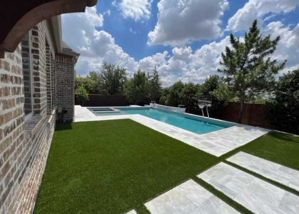 Modern backyard with artificial grass, rectangular swimming pool, and white stone pathway beside a brick wall, under a clear blue sky.